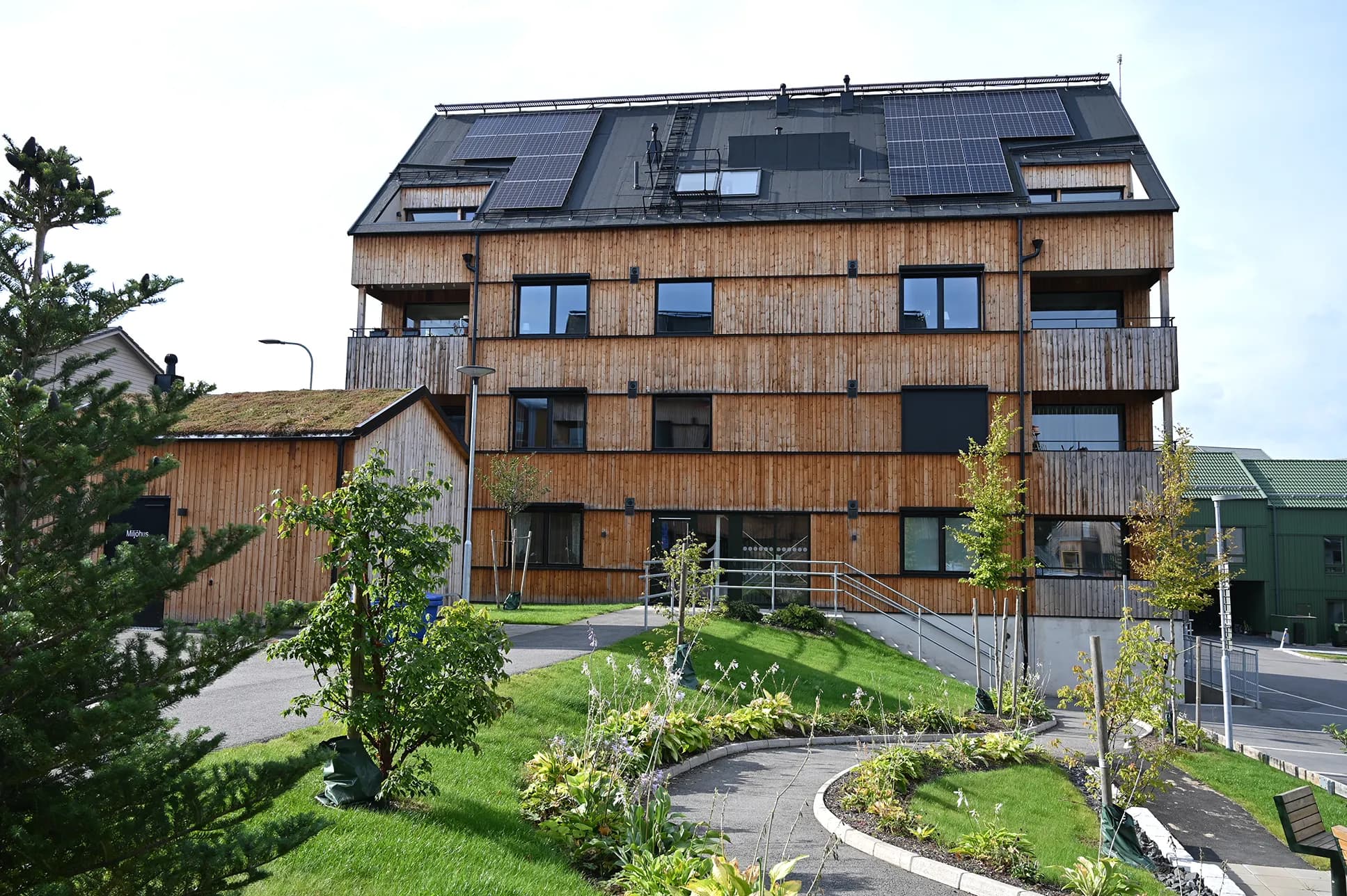 Flerbostadshus i trä med balkonger och solpaneler på taket, omgivet av grön gård med slingriga gångvägar